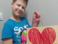 Ósme zdjęcie przedstawia zadowolonego Jakuba Wiewiórę, trzymającego w  dłoni drewnianą deseczkę na której zostało wykonane czerwone serce metodą string artu, która polega na nabijaniu gwoździkami wzoru a następnie przewlekanie pomiędzy nimi kolorowego sznurka.